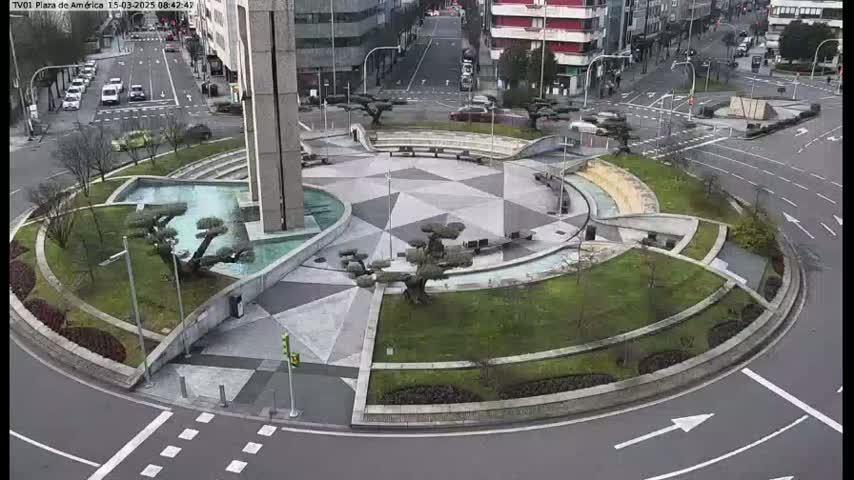 An aerial view shows a circular plaza with landscaped areas, a geometric patterned pavement, and a water feature, surrounded by roadways with light traffic on an overcast day.