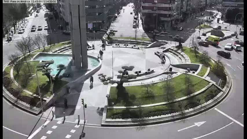 A circular plaza with a fountain, landscaping, and seating areas is seen from an overhead perspective on a sunny day, with cars visible on surrounding streets.