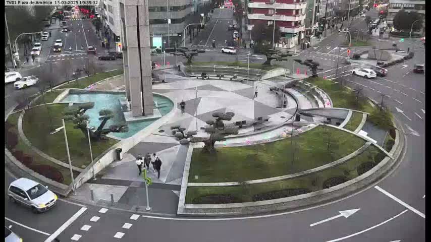 A circular plaza with a central fountain and landscaping, surrounded by roads with cars and pedestrians, on an overcast day.