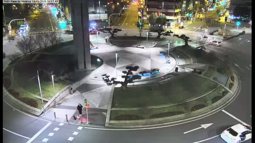 A nighttime aerial view shows a circular plaza with landscaped areas and small trees, surrounded by a roadway with a few cars visible under artificial light.