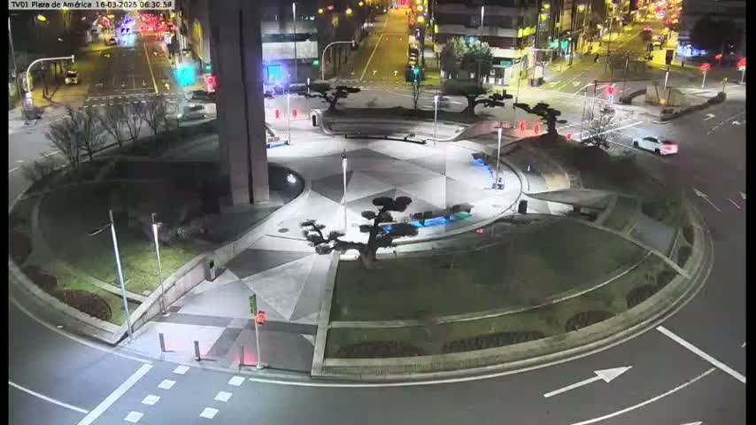 A nighttime aerial view shows a circular plaza with landscaped areas, geometric pavement designs, and low-lying vegetation, situated at a roadway intersection.