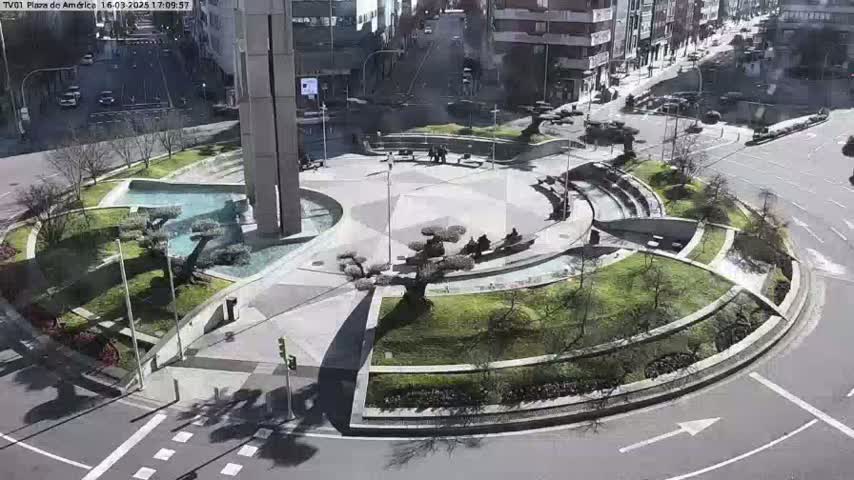 A sunny day reveals a circular plaza with a central fountain, landscaped areas, and surrounding buildings, intersected by roadways.
