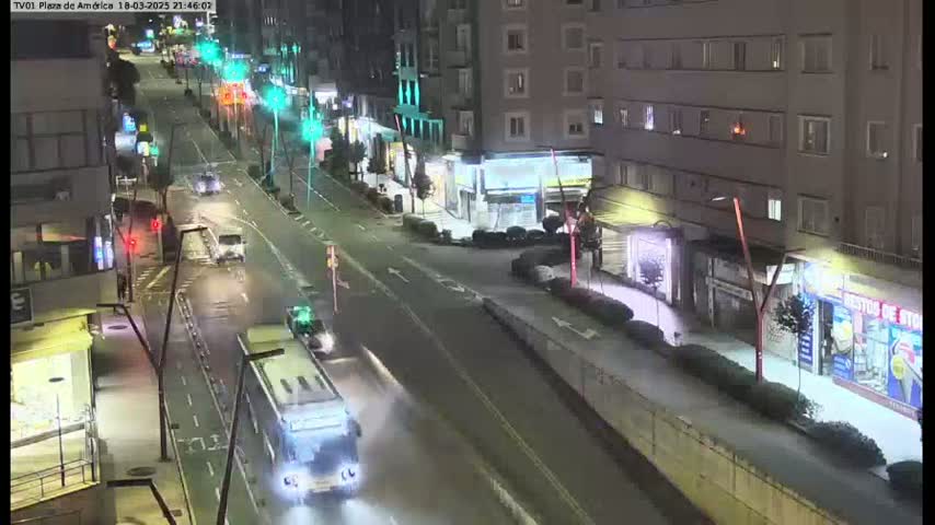 A wet, nighttime street scene shows several vehicles traveling along a city road lined with buildings and shops.