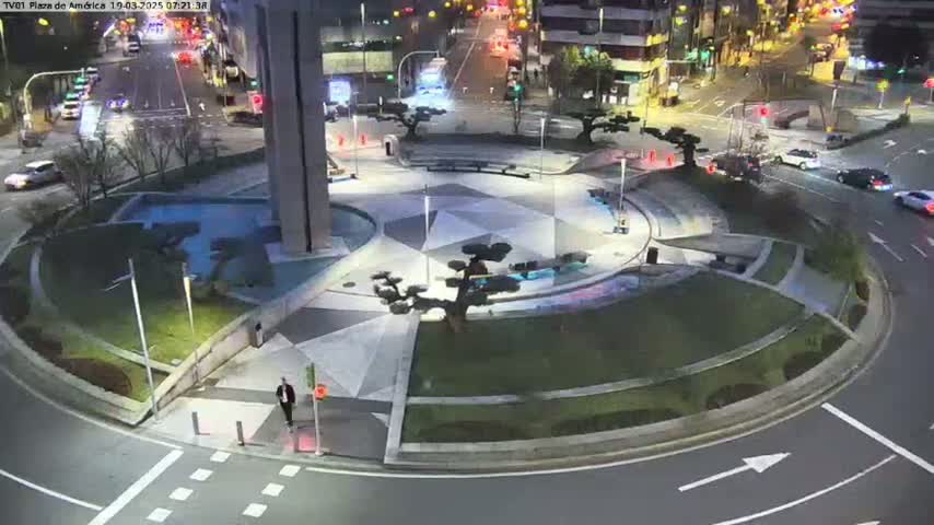A nighttime aerial view of a circular plaza with geometric landscaping, surrounded by roadways with sparse traffic.