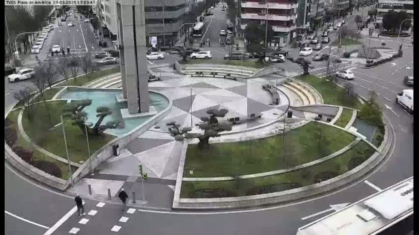 A roundabout in a city with a geometrically designed central plaza featuring landscaping and water features, surrounded by roads with cars and pedestrians.