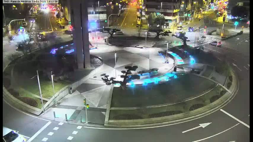 An illuminated roundabout at night features geometric paving, artificial trees, and blue-lit water features, surrounded by roadways with minimal traffic.