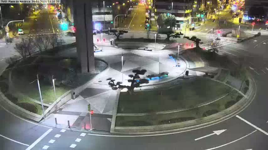 An illuminated plaza at night features geometric landscaping, small trees, and a roundabout with surrounding roadways.