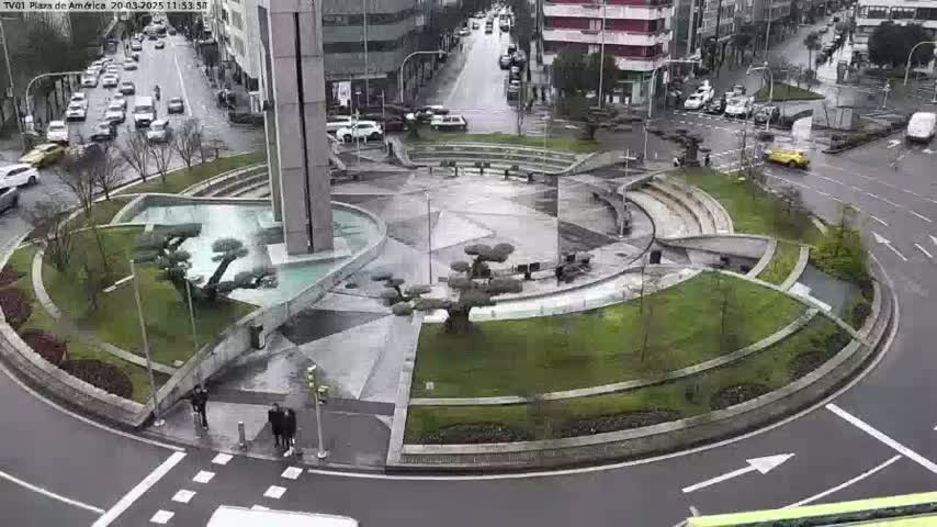 A circular plaza with landscaped areas and a central fountain is surrounded by a roadway with cars driving on it in rainy weather.