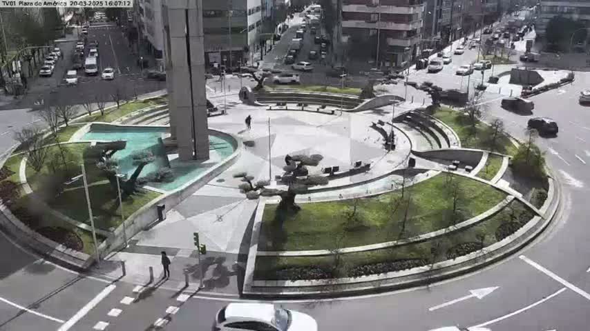 A circular plaza with a fountain and landscaping is surrounded by streets with cars driving on a sunny day.