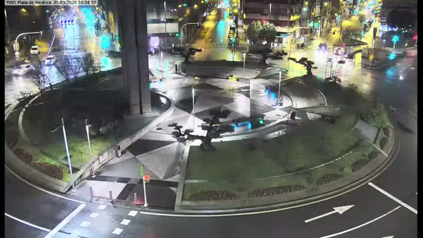 A nighttime view of a circular plaza with a geometric design, featuring several small trees and minimal traffic on wet roads.