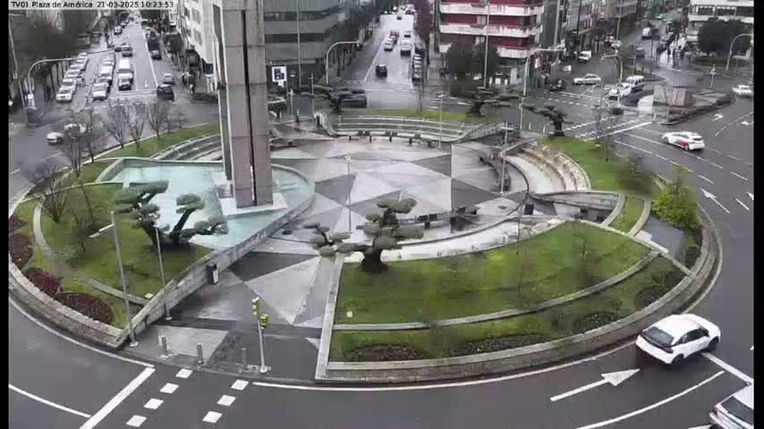 A landscaped traffic circle with a central plaza, surrounded by buildings, and a few cars on a wet, overcast day.