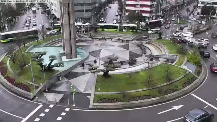 A circular plaza with geometric landscaping and a central water feature is surrounded by a roadway with cars and buses, all under a rainy sky.