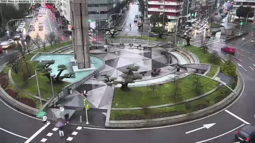 A rain-slicked roundabout in a city center features a geometric plaza with a central fountain and several small, manicured trees, surrounded by roadways with cars and pedestrians.