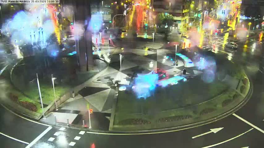 A rain-slicked roundabout at night features a central plaza with colorful water features and sparse vehicle traffic.