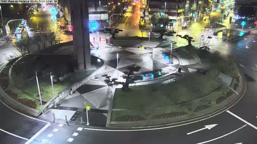 A nighttime, wet roundabout with geometric landscaping and sparse, low shrubbery is illuminated by streetlights, and some traffic is visible on the surrounding roads.