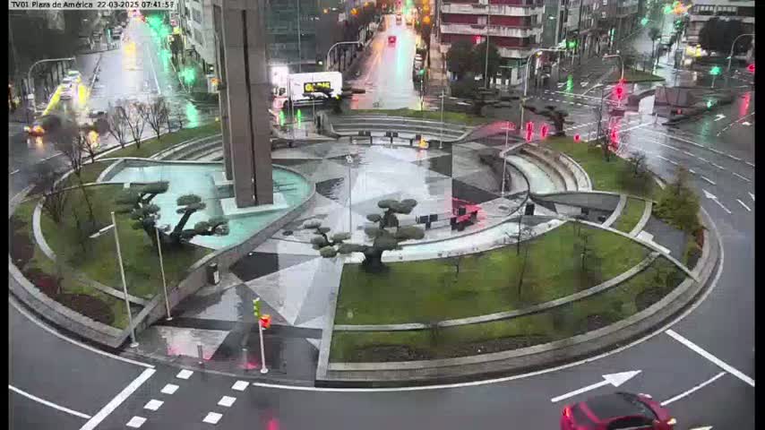 A wet, circular plaza with landscaping and a fountain is situated in the center of a roundabout, surrounded by roadways with traffic.