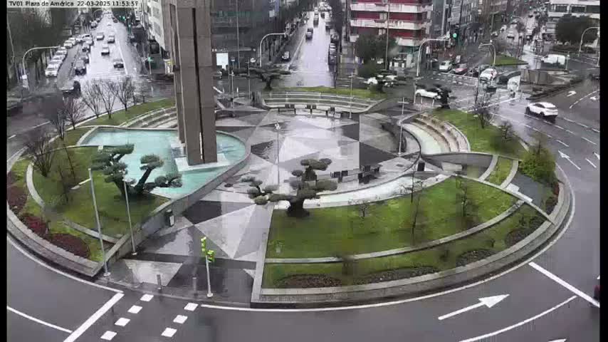 A rain-slicked roundabout features a geometrically designed plaza with landscaping, a central fountain, and several small trees.