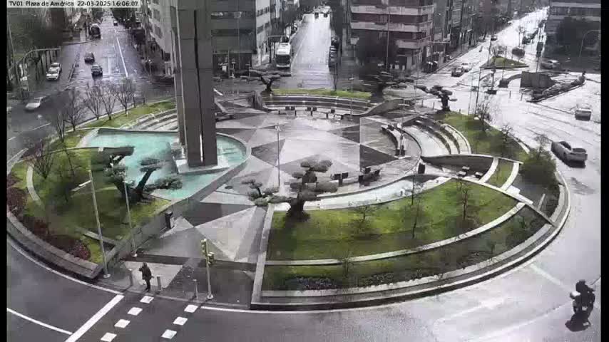 A wet, geometrically designed plaza with a central fountain and landscaping is surrounded by a circular road with sparse traffic under a rainy sky.