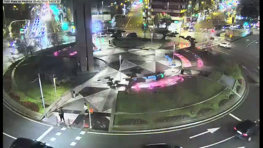 A nighttime aerial view of a roundabout with a geometrically patterned center, surrounded by green landscaping and with cars visible on the roads.