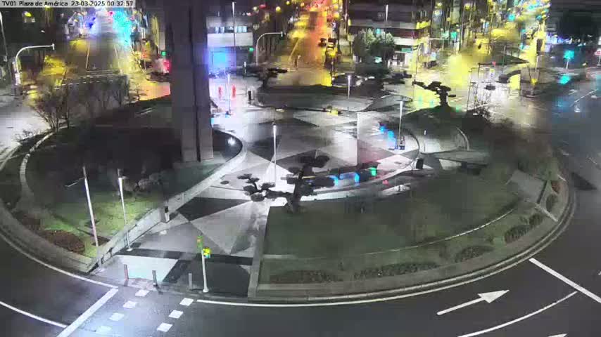 A nighttime aerial view of a roundabout with geometric landscaping and sparse traffic, under wet conditions.