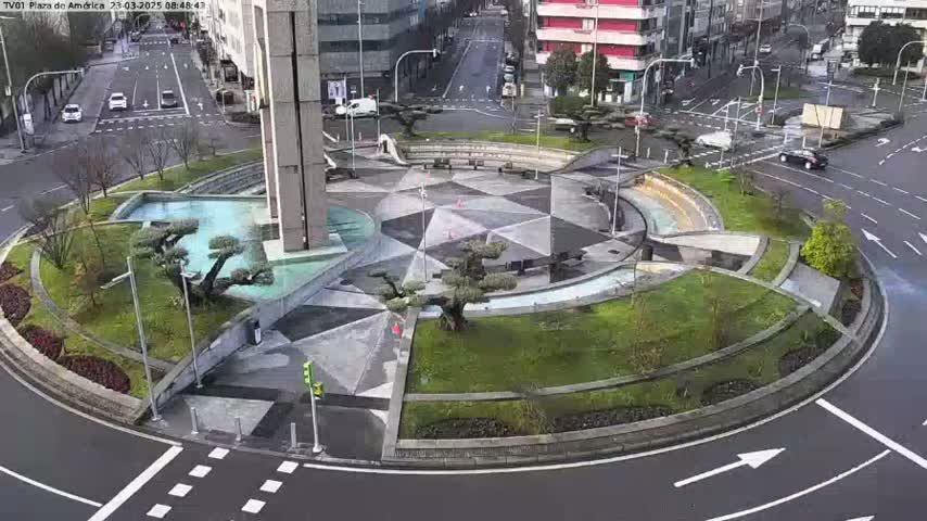 An aerial view shows a geometrically designed roundabout with a central plaza featuring landscaping and small water features, surrounded by city streets and buildings on an overcast day.