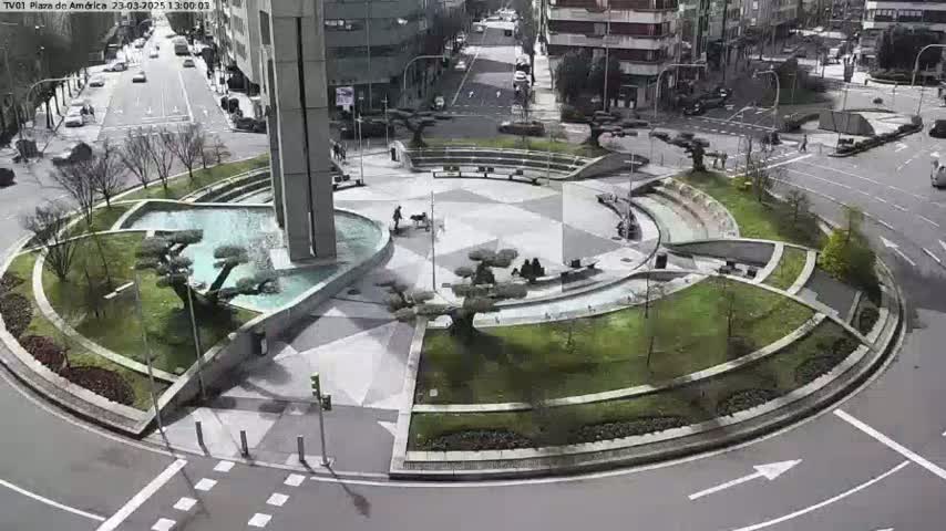A circular plaza with a fountain, landscaping, and several people is situated at a roadway intersection on a sunny day.