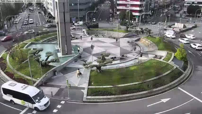 A high-angle view of a roundabout with landscaped areas and a water feature, surrounded by streets with cars and pedestrians, on a partly sunny day.
