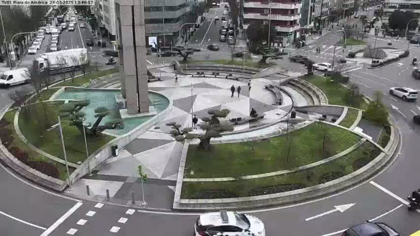 A circular plaza with landscaped areas and a central fountain is surrounded by a roadway with cars, under an overcast sky.
