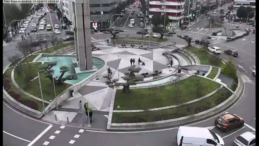 A circular plaza with a fountain and landscaping is surrounded by roads with cars and pedestrians on an overcast day.