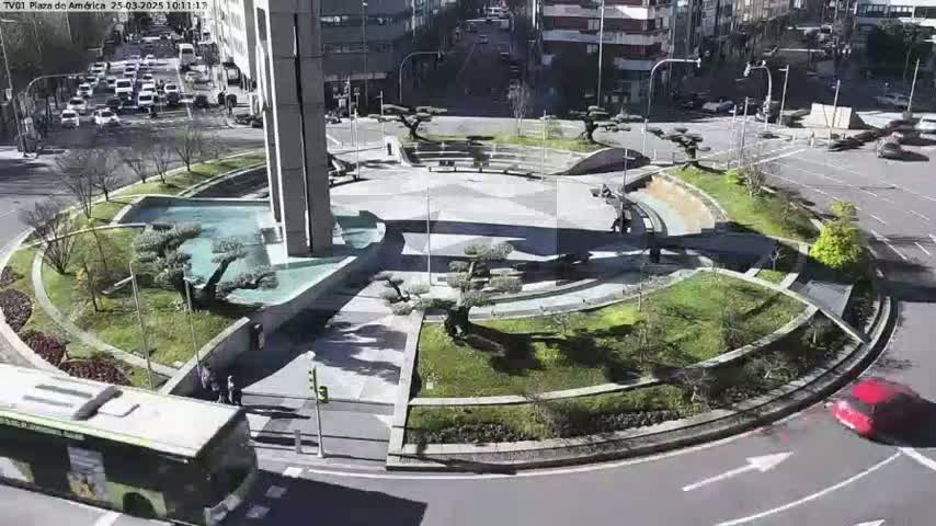 A sunny day reveals a circular plaza with landscaped gardens, a central fountain, and a nearby street with cars and a bus.