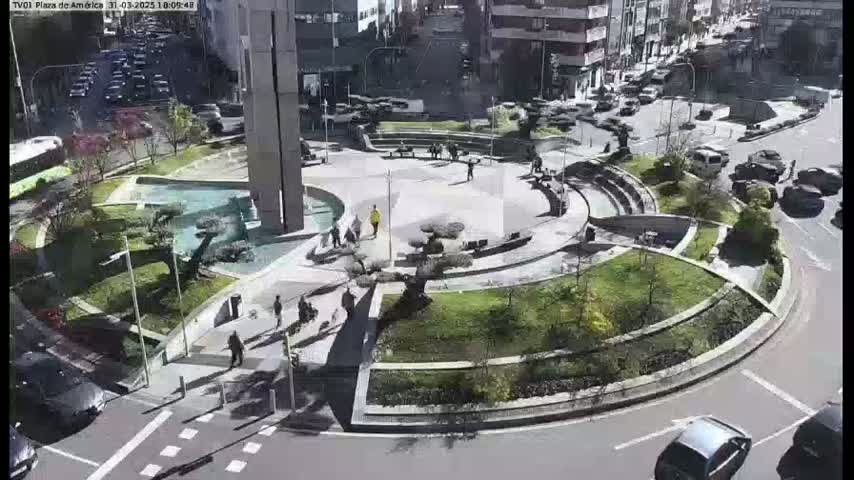 A circular plaza with landscaped gardens and a fountain is surrounded by city streets with cars and pedestrians on a sunny day.