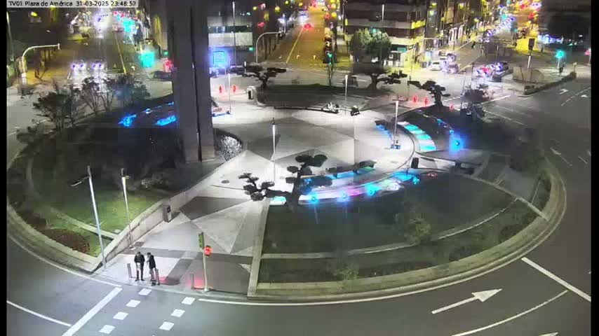 A nighttime aerial view of a roundabout with illuminated landscaping features and two pedestrians standing near a crosswalk.