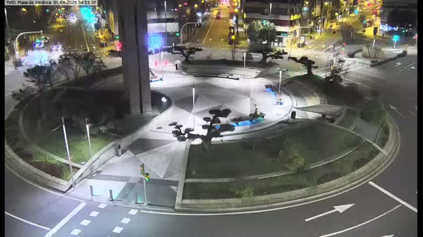 A nighttime aerial view shows a circular plaza with geometric landscaping, small trees, and illuminated pathways, situated within a larger roadway intersection.