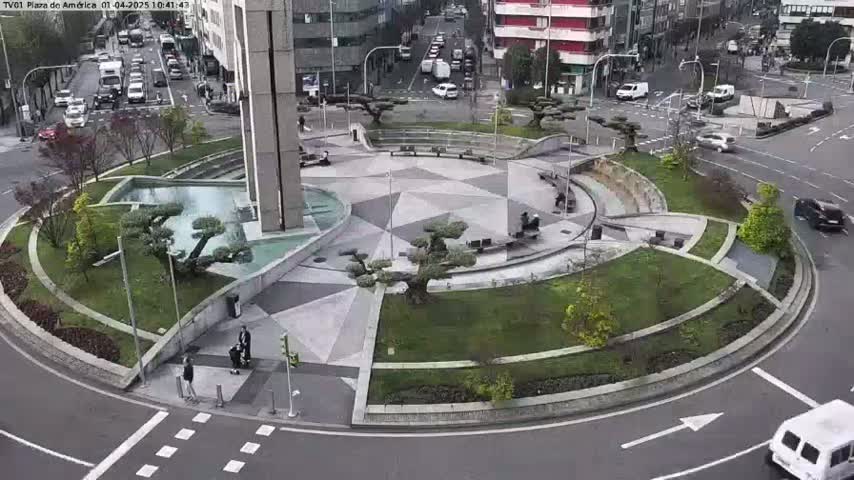A landscaped roundabout with a geometric pattern in the center, surrounded by a roadway with a few cars and pedestrians, on a partly cloudy day.