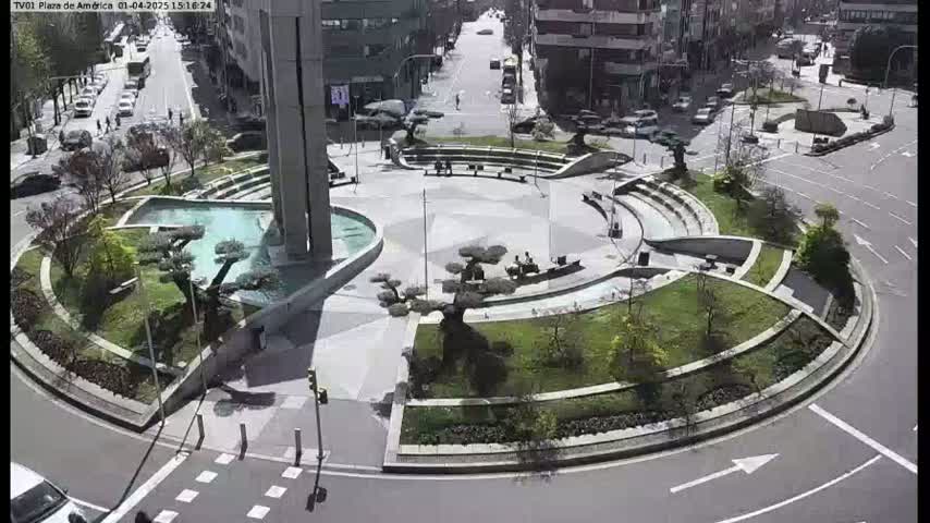 A circular plaza with a fountain, landscaping, and seating areas is surrounded by roadways on a sunny day.