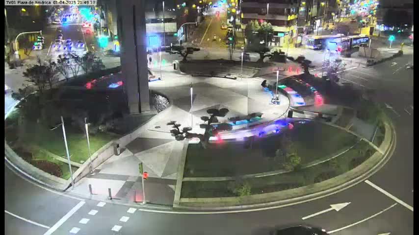 A nighttime aerial view shows a circular plaza with geometric patterns and landscaping, surrounded by roads with cars and a bus, under clear skies.