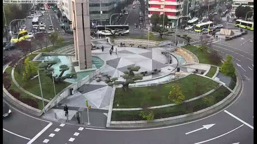 A circular plaza with geometric landscaping, water features, and small trees is situated in the center of a roundabout, with vehicles and pedestrians visible on surrounding streets under an overcast sky.