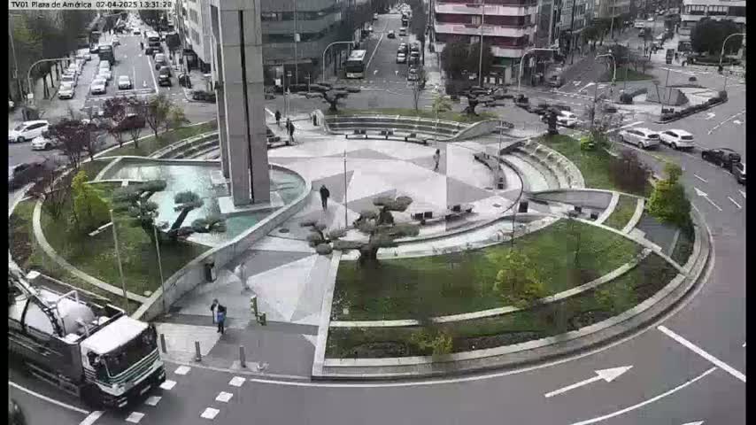 A cement roundabout with landscaping features, including small trees and a water fountain, is surrounded by city streets with cars and buildings in the background.
