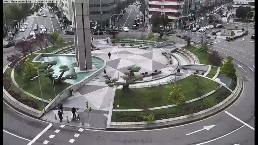An aerial view shows a circular plaza with a geometric pattern, landscaping, and a fountain, surrounded by a roadway with cars; the weather appears overcast.