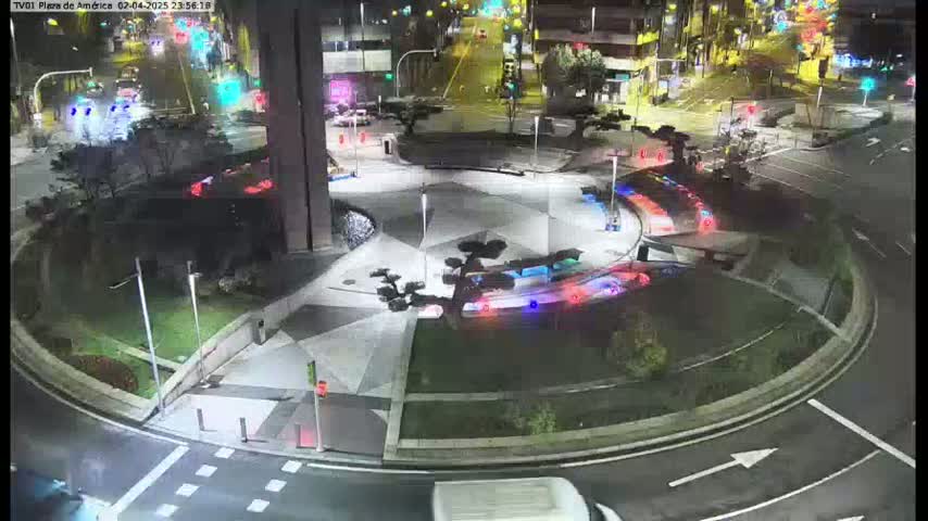 A nighttime view of a roundabout with a geometrically patterned center, surrounded by landscaping, with several cars visible on the roads leading to and from it.