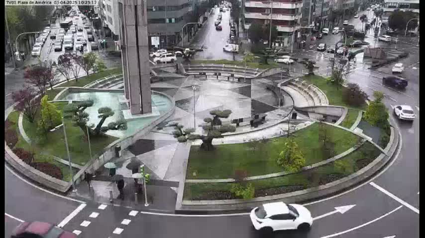A rain-soaked roundabout features a central plaza with water features and manicured landscaping, surrounded by a roadway with several cars.