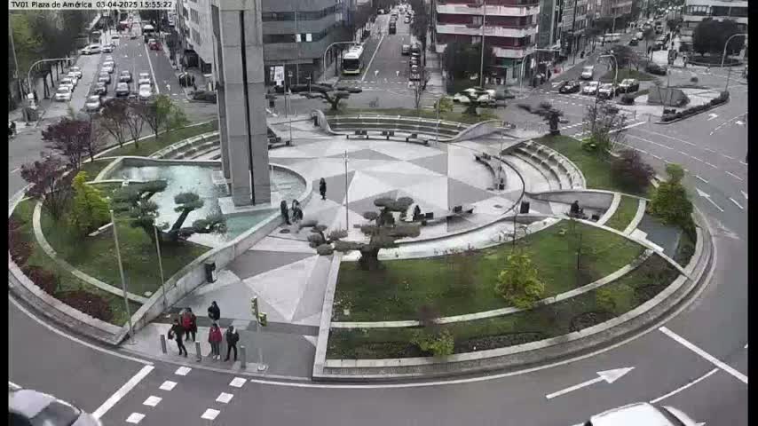 A circular plaza with a central fountain, landscaped areas, and seating is surrounded by a roadway with cars, under an overcast sky.