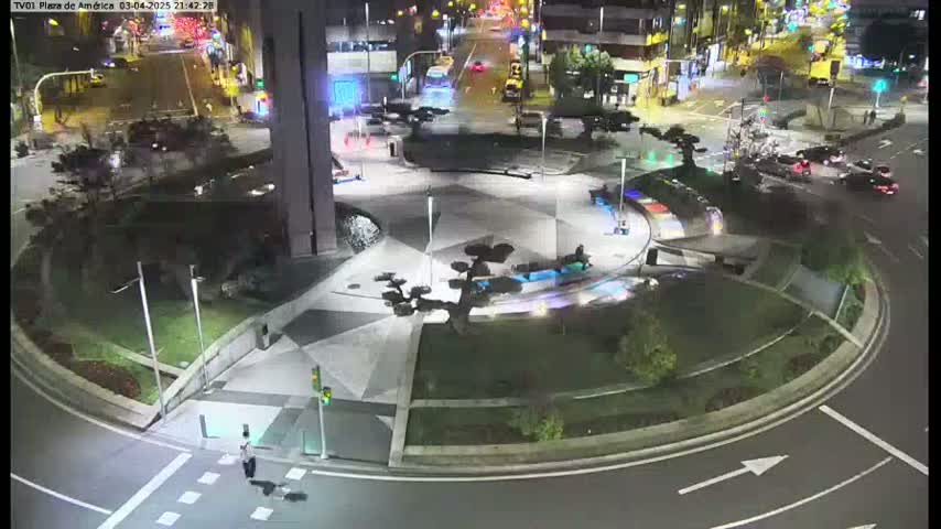 A nighttime aerial view of a plaza with geometric landscaping, featuring small trees and a circular roadway, with a few cars visible on the surrounding streets.
