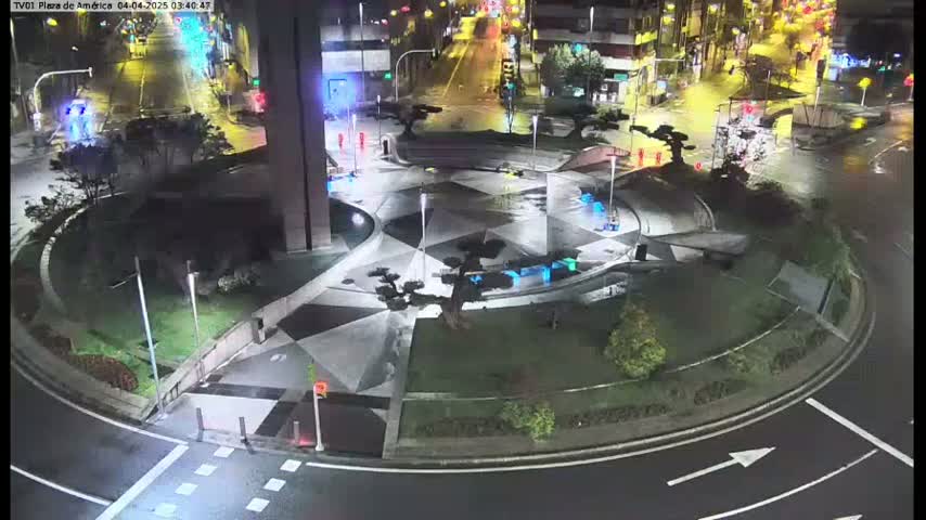 A nighttime aerial view of a roundabout with a geometrically patterned plaza in the center, surrounded by wet roads and landscaping.