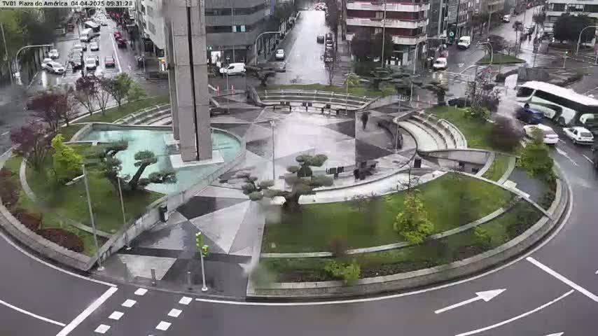A roundabout landscaped with shrubs and small trees, featuring a central water feature, is surrounded by city streets with light traffic in rainy weather.
