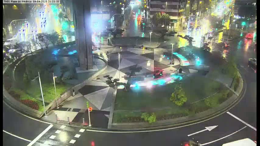 A wet, nighttime view of a roundabout with a geometrically patterned plaza in the center, featuring illuminated fountains and sparse vehicle traffic.