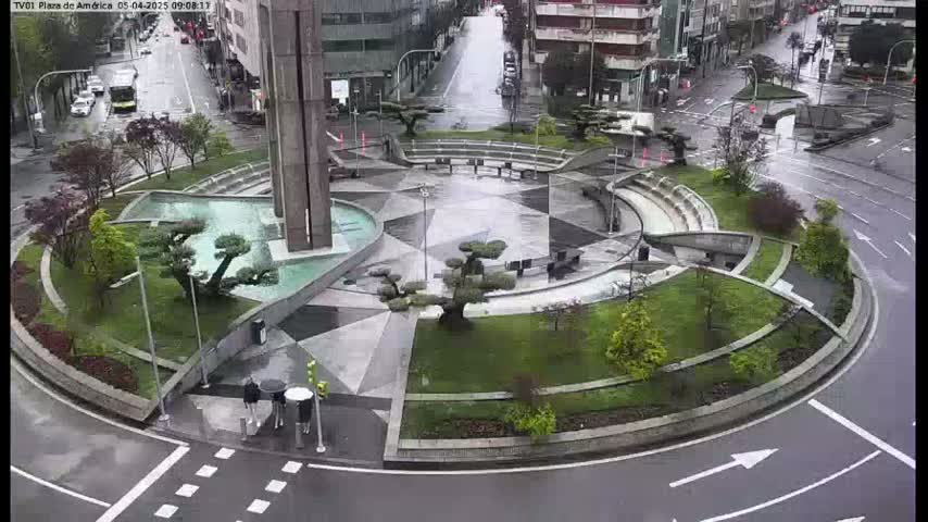 A rain-slicked roundabout features a modern, geometric plaza with landscaped gardens and small trees.