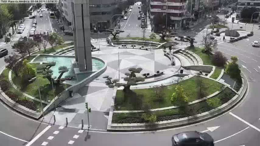 A circular plaza with a fountain, landscaping, and seating areas is surrounded by roadways with cars and buildings in the background on a sunny day.