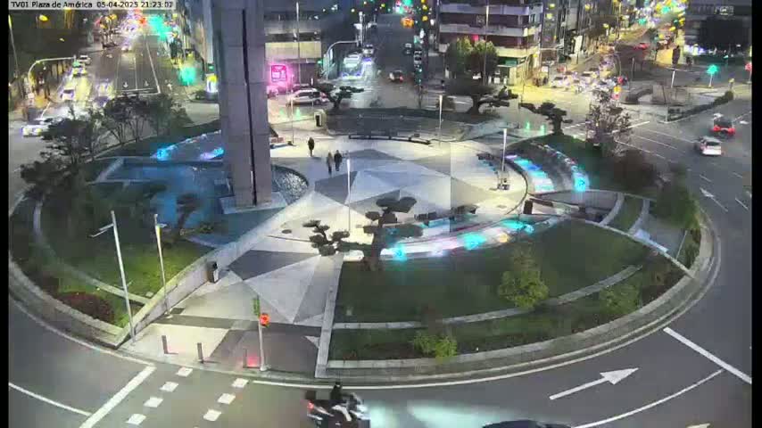 A nighttime aerial view of a roundabout with a geometrically patterned plaza featuring illuminated water features, surrounded by roadways with sparse traffic and low-level lighting.