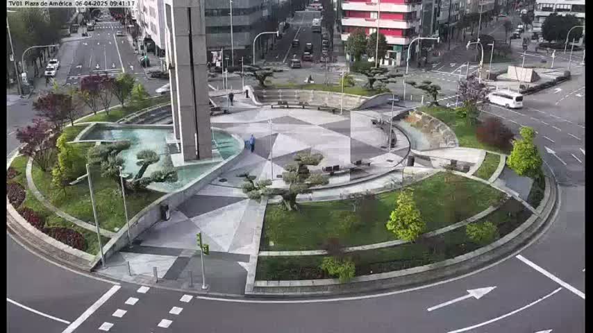A landscaped traffic circle with a geometrically patterned center, featuring water features and several small trees, is surrounded by a roadway on a partly cloudy day.